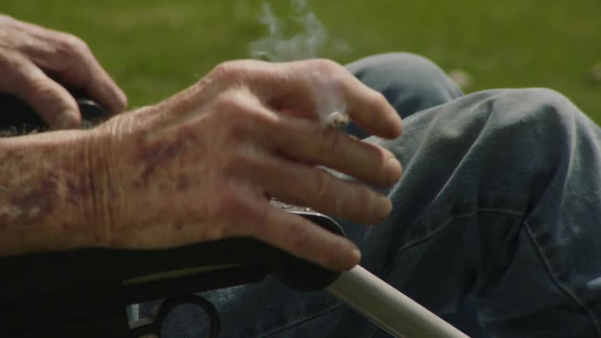 A closeup of an elderly persons hands as they hold and flick the ash from a cigarette while resting on a chair arm