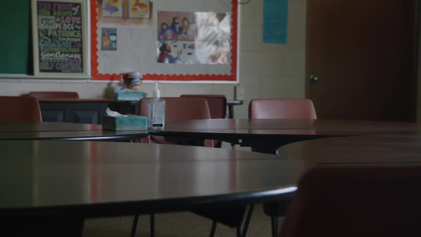 The video shows an empty classroom with a large table and several chairs the room is dimly lit with light coming in from a window on the far side on the wall theres a bulletin board and a blackboard and in the corner a bookshelf is visible
