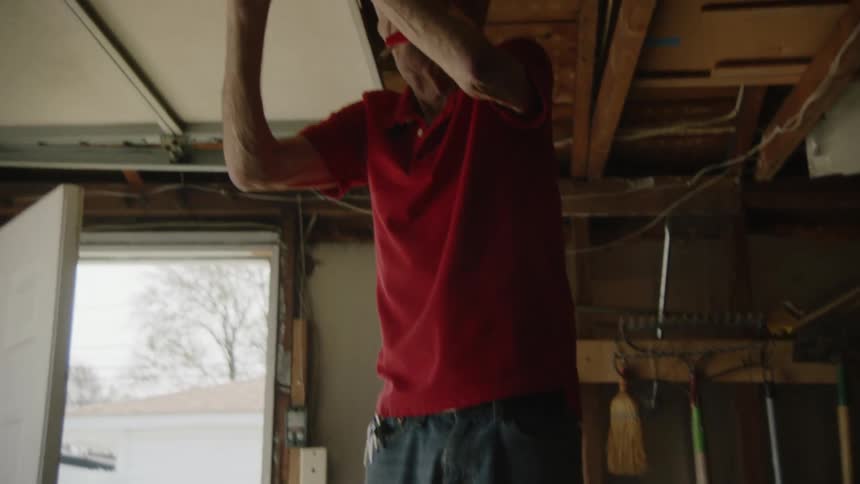 A man wearing a red shirt blue jeans and black shoes stands on the top step of a small metal ladder reaching up with both hands to install a light fixture in a garage with a white bucket and various tools visible against the far wall