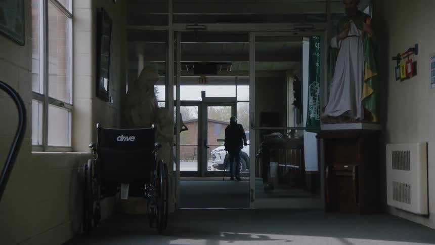 A shot from inside a building shows a person walking toward a glass door the person is holding a cane or walking stick a wheelchair is visible in the foreground
