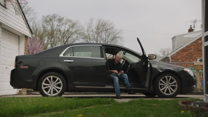 The video shows an elderly man sitting in the drivers seat of a black car parked in a driveway he is looking down and appears to be putting on his shoes before exiting the car a white garage and a house with a large window are visible in the backgroun