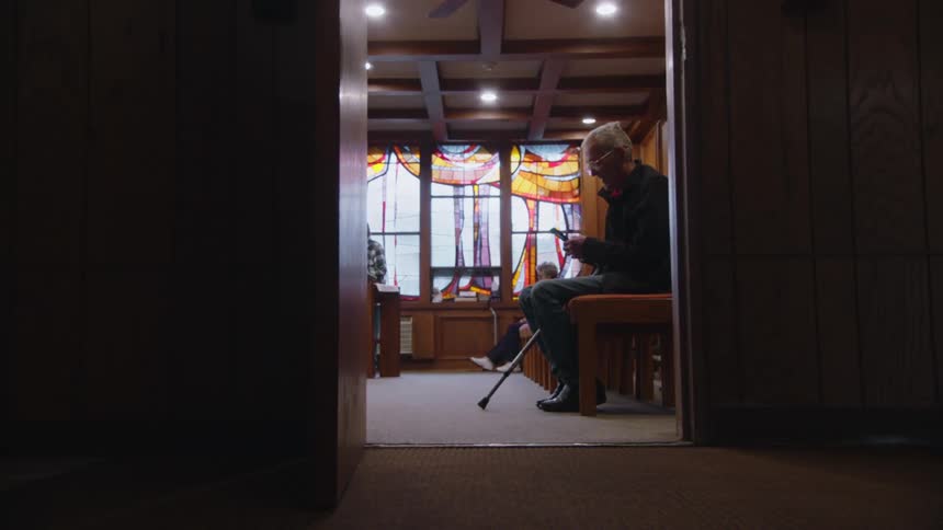 Elderly man sitting on a bench with a cane looks at his phone in front of a colorful stained glass