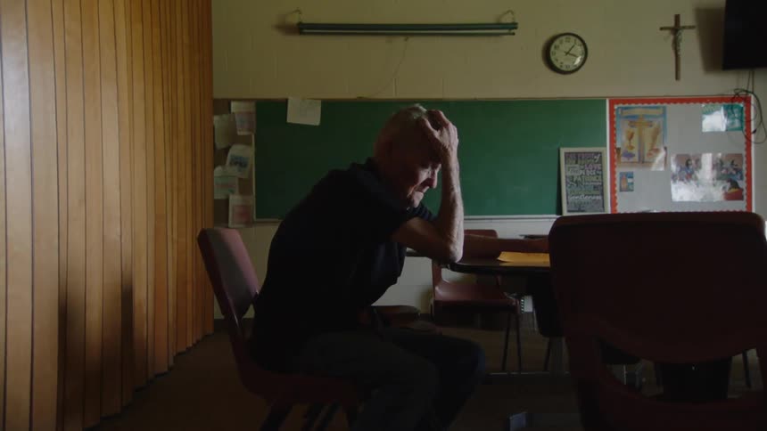 A elderly man sits alone in a dimly lit classroom in a moment of distress