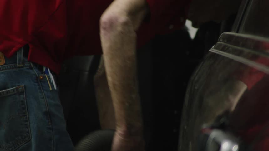 An elderly man with gray hair in a red shirt and jeans as he bends down and uses a vacuum cleaner to clean the interior of a car