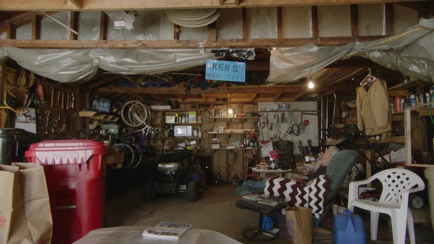 The video shows the interior of a cluttered workshop or garage with various tools boxes and a bicycle hanging on the walls in the middle of the room there are several chairs and a sign on the wall that reads kens man cave