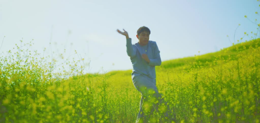 A man wearing a blue top and pants dances in a field of tall yellow flowers with a bright sunlit sky and a green hill in the background