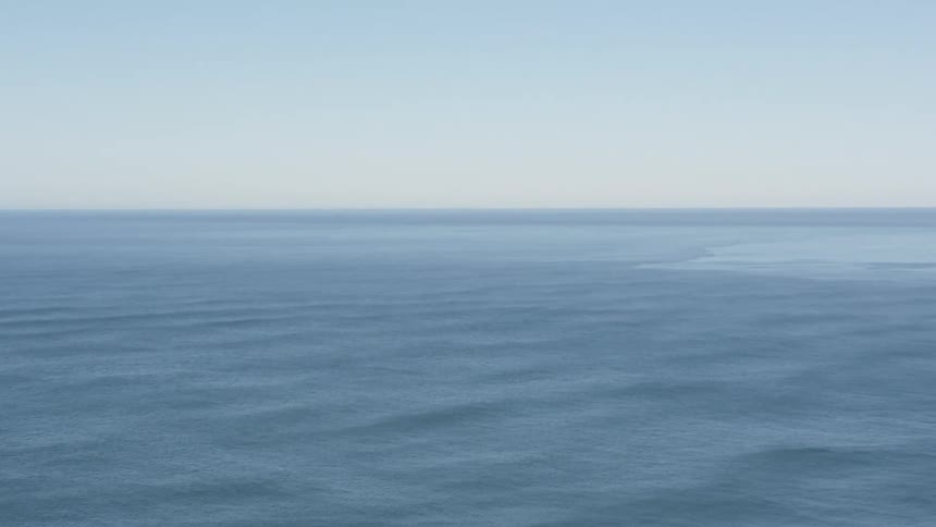 The video shows a static wide shot of the vast ocean on a clear day the calm blue water extends to the horizon where it meets a lighter blue almost white sky creating a serene and minimalist seascape
