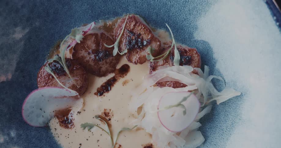 The video shows a detailed closeup of a gourmet dish served in a blue bowl the meal consists of seared scallops on a creamy sauce garnished with thinly sliced radishes and delicate green sprouts creating an appetizing and elegant presentation