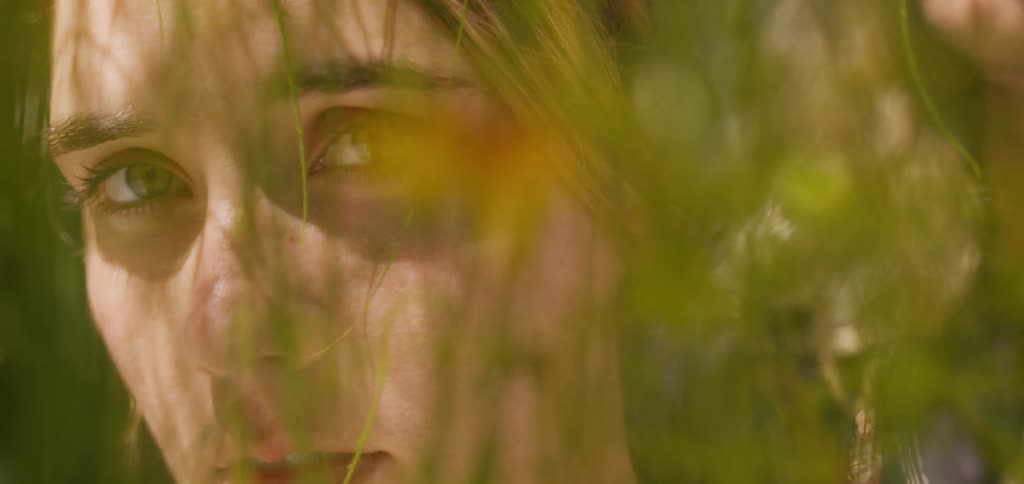The video shows an extreme closeup of a womans face the lighting creates a striking contrast with one side of her face in bright sunlight and the other in deep shadow while a thin plant stem lightly rests on her cheek