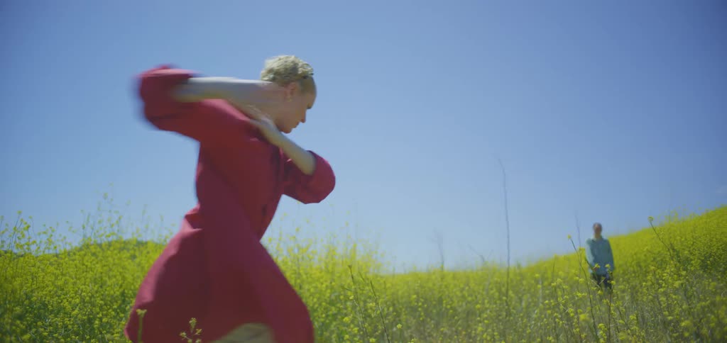 Woman in a pink shirt dancing in a field of yellow flowers and another person in the background