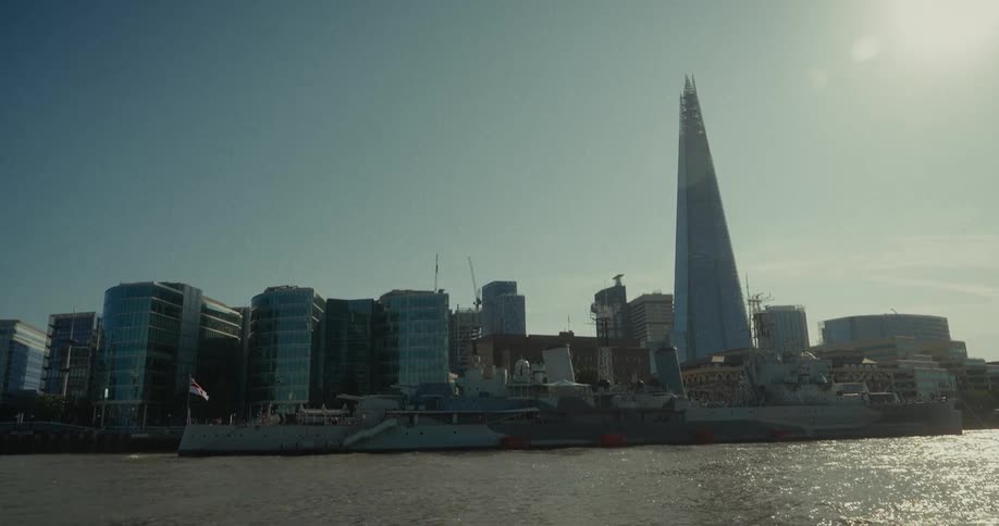 A long view from behind a metal railing shows a large ship docked on a river with a city skyline in the background featuring several tall buildings and a prominent glass skyscraper