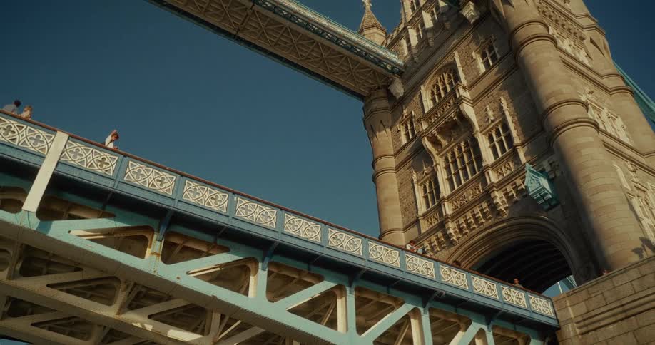 A high angle upward looking view shows the underside of londons tower bridge revealing its intricate gray steel structure as the camera pans up the side of a tall stone tower with people walking on the upper level