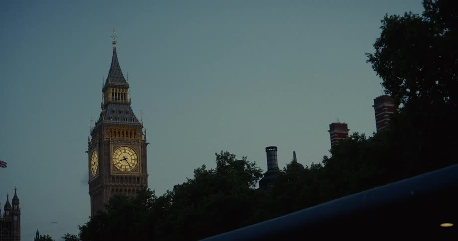 The illuminated face of the big ben clock tower is seen against the twilight sky with treetops and chimneys in the foreground