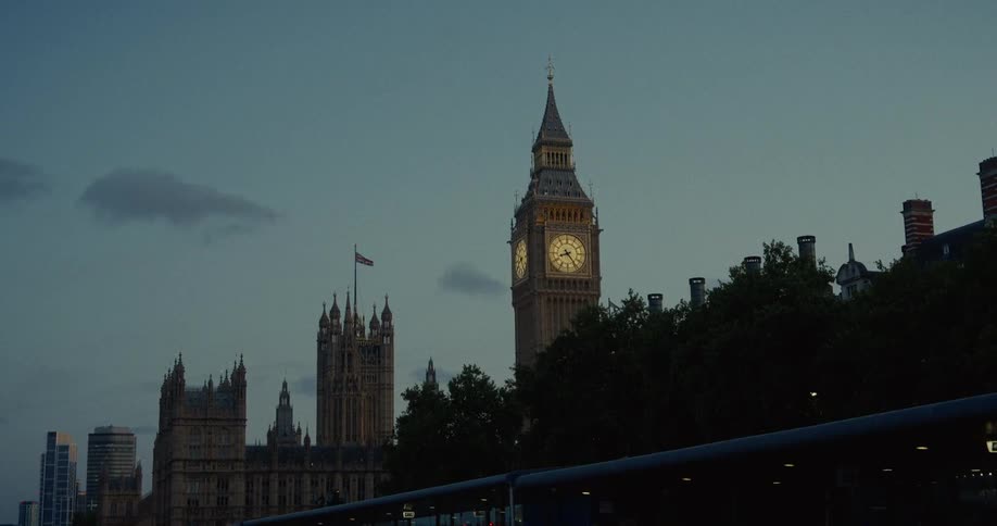 A long view shows londons big ben clock tower with its faces lit up in a pale yellow standing beside the shadowed houses of parliament and a distant skyscraper under a cloudy evening sky