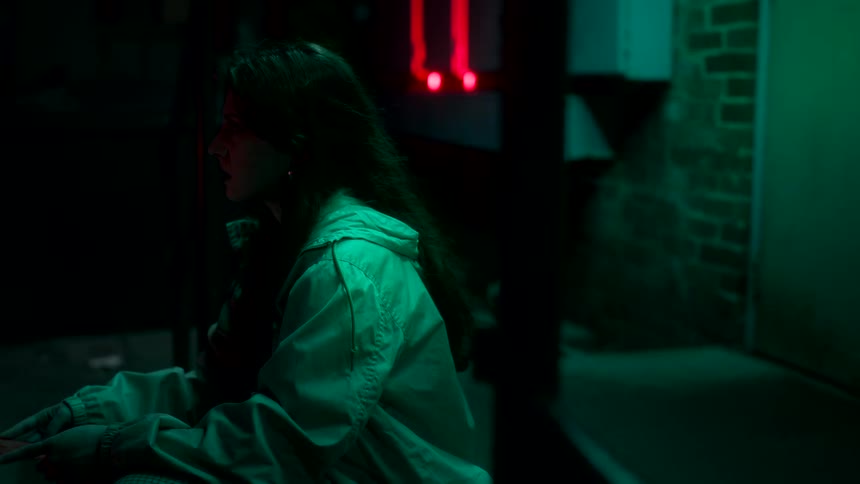 A young woman converses and contemplates while holding a card as she sits in front of a building at night