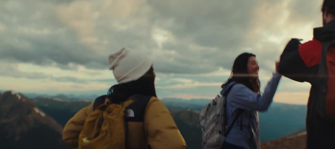 Three people in warm clothes are on top of a mountain and are clapping their hands in greeting on a day with many clouds in the sky