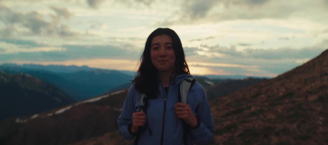 Woman with a backpack smiles gently in front of mountains at sunset