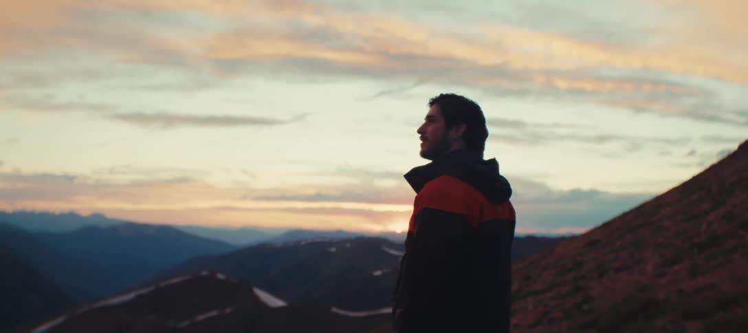 Man in a black and red jacket gazes at the horizon during sunset in the mountains