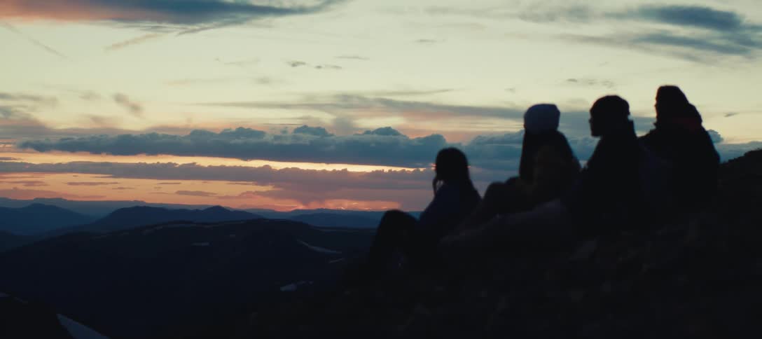 A group of four friends sit outdoors on a mountain slope and watch the sunset