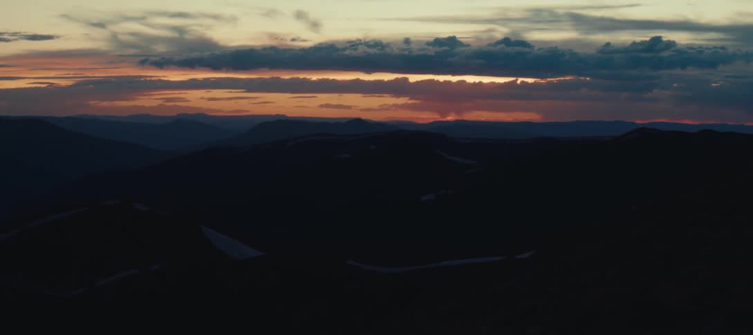 A landscape of a dark mountain range in the morning with the sun shining in the sky turning everything orange