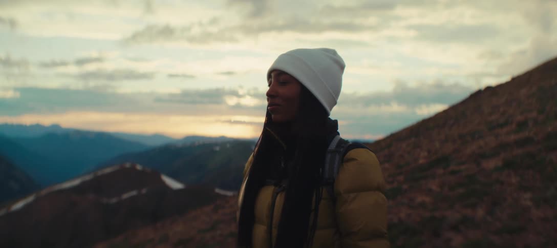 Woman in a white beanie and yellow jacket gazes at the mountain landscape at dusk