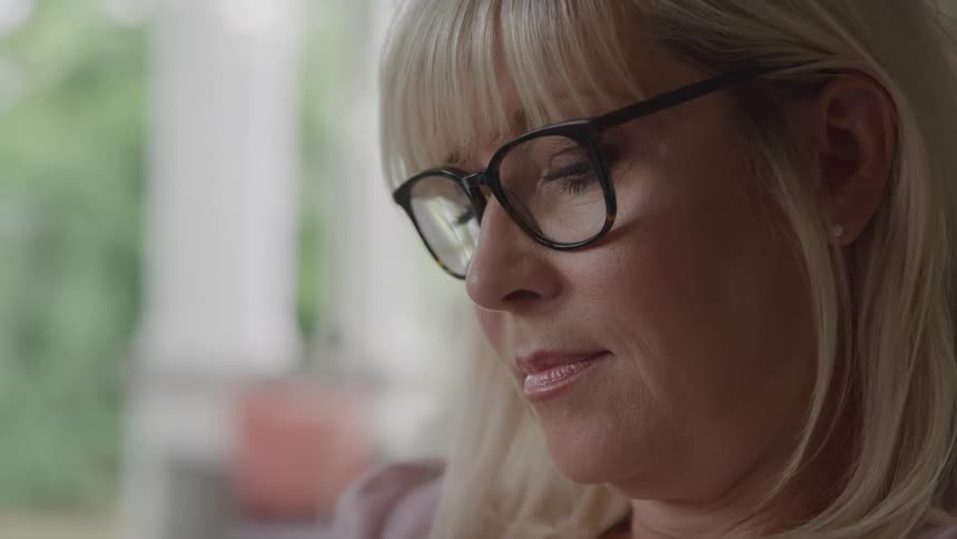 The face of a blonde woman wearing blackframed glasses and looking down indoors