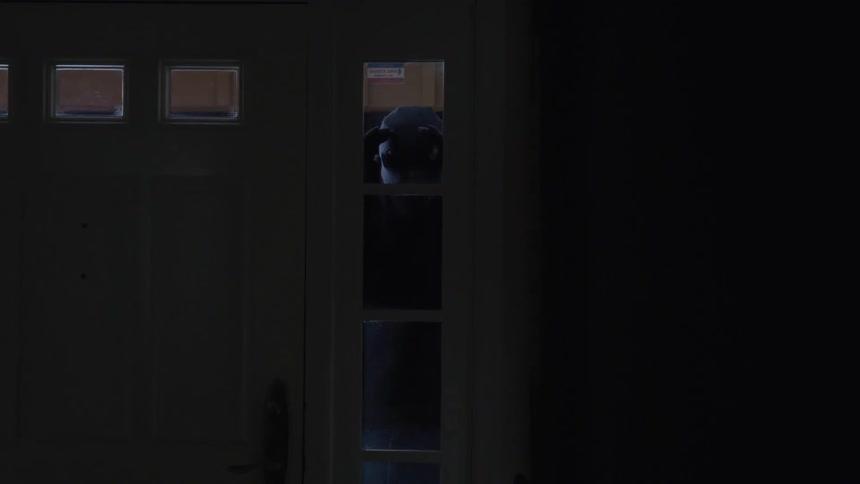 A man wearing a mask and gloves looks in a window next to a door before breaking in