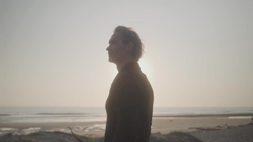 A silhouetted person is standing on a beach facing the ocean the sun is behind them casting a bright light and creating a halolike effect on their hair