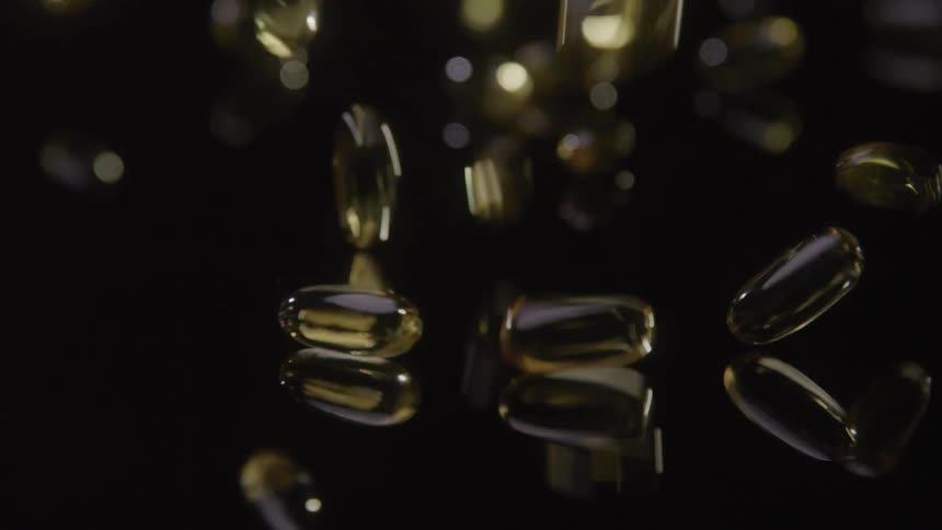 A close up shows several yellow oblong gelatin capsules falling onto a reflective black surface bouncing before coming to a rest