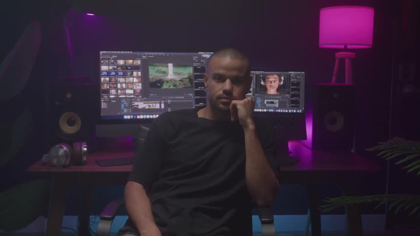 A young black man sits in front of a work desk with computers in a music studio