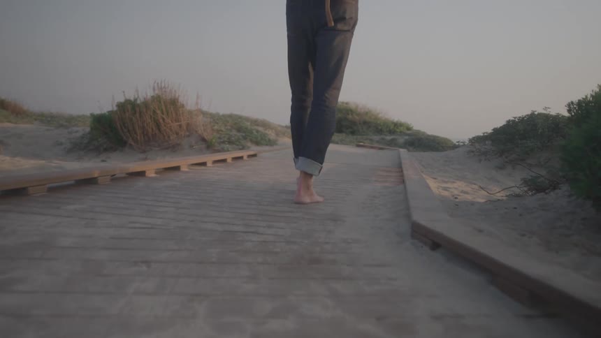 A closeup shot shows a persons bare feet walking on a wooden boardwalk the sun is setting behind them casting a soft golden glow on the sandy beach around the walkway