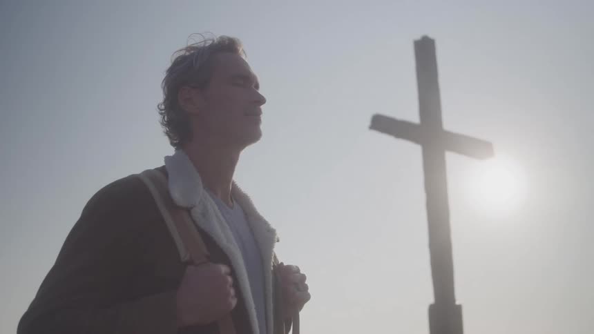 A man with a backpack on his back stands looking to the side with a calm expression as a large wooden cross stands in silhouette against the bright sun