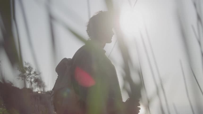 A silhouette of a man with a backpack walks to the right across the frame with long grass obscuring the bottom half of the image and the bright sun creating a flare in the background