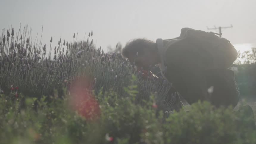 Man carrying a backpack kneels to smell the flowers growing outdoors on a cloudy day