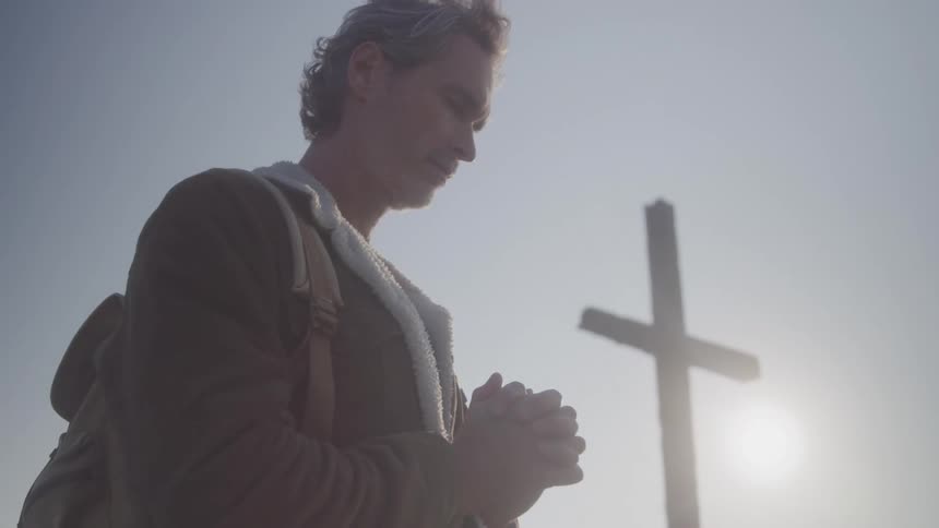 Man wearing a jacket and carrying a backpack prays near a large cross on a sunny day