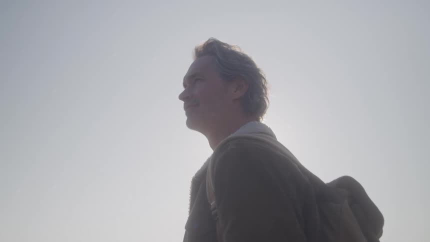 Man with a backpack looks ahead under a clear sunlit sky