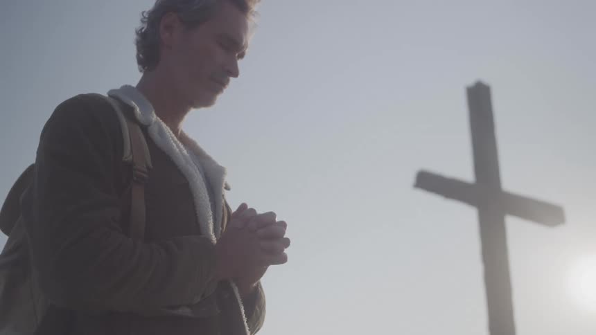 Man with closed eyes prays in front of a wooden cross outdoors