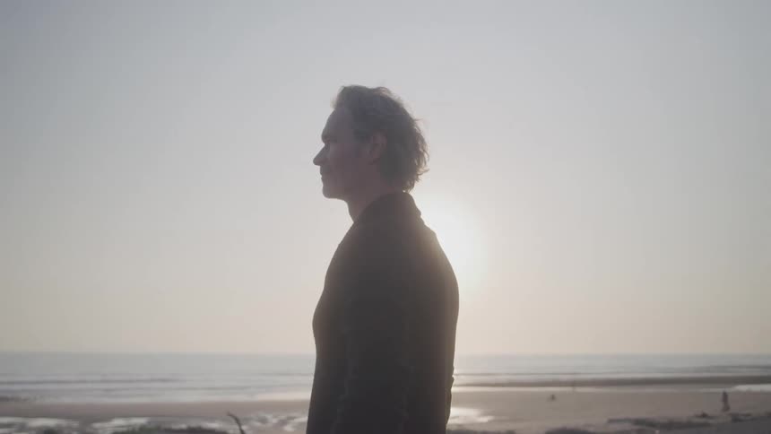 A silhouetted figure with graying hair stands on a beach facing the ocean the person is backlit by a bright hazy sun creating a strong rim light effect around their head