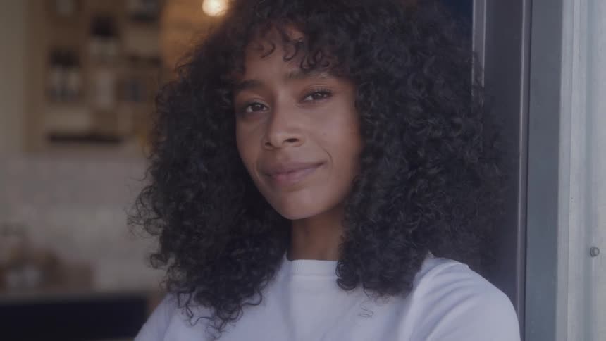 A young woman with dark skin and curly black hair smiles as she stands in a doorway looking directly at the camera
