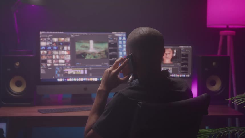 A man in a chair facing away from the camera intensely focused on his computer monitors as he works on a video editing project and talks on a smartphone
