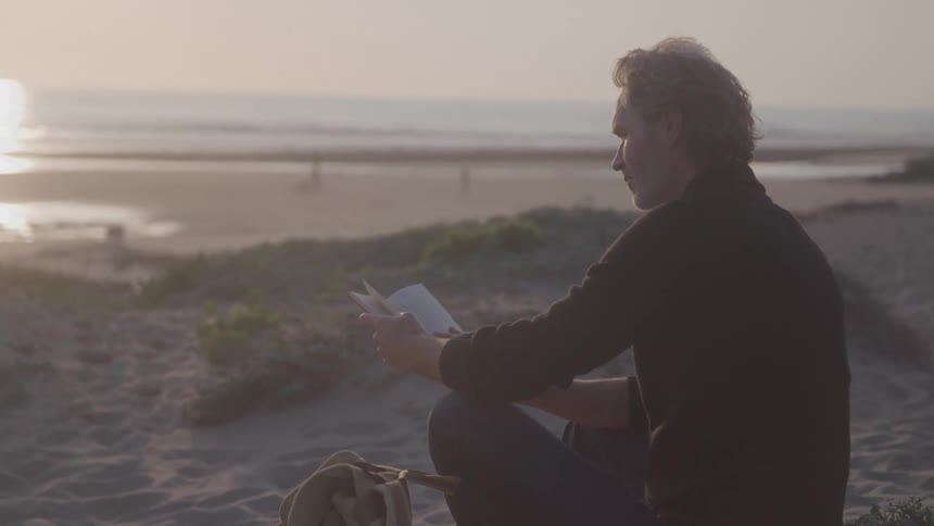 The man is reading while sitting on the sand