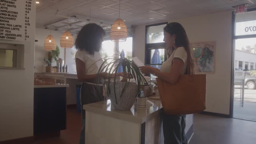 A young waitress with dark curly hair stands behind a counter and smiles while interacting with a customer