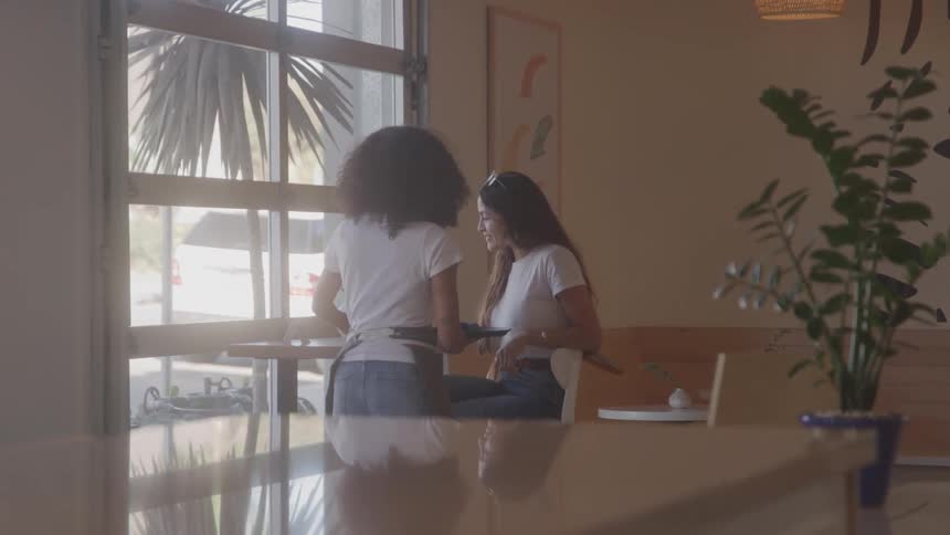 A steady shot of a woman with long dark hair sitting alone at a small table in a modern cafe while another woman is serving her some coffee