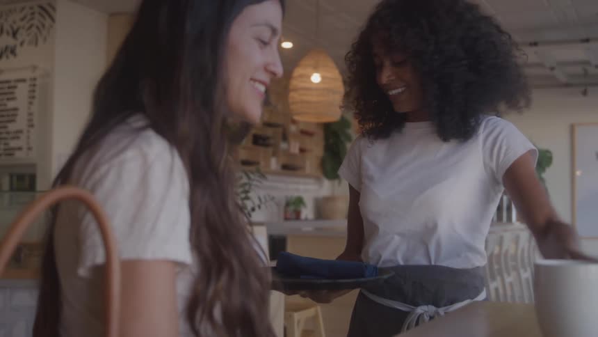 A waitress with curly black hair stands behind a counter and smiles while carrying a tray with two white mugs to a customer with long brown hair who sits at the counter holding a cell phone