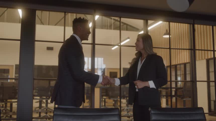 Two professionals in suits shake hands in a glasswalled meeting room