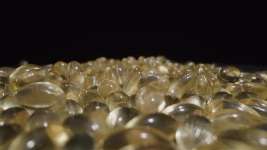 A macro camera shot slowly pushes in on a large pile of shiny goldencolored soft gel capsules against a dark background