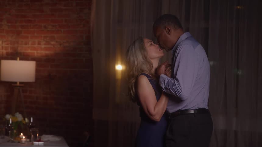A middleaged couple in formal attire slow dances together in a dimly lit intimate room with a rustic brick wall