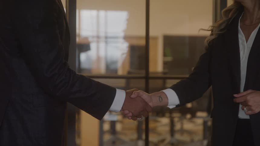 A man and a woman standing in an office near a glass give each other a handshake while another woman walks by on the other side of the glass