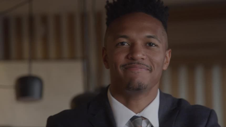 A closeup of a man in a dark suit with a tie smiling confidently and making eye contact with the camera
