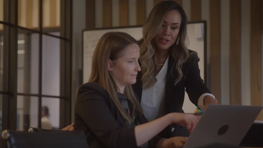 Two coworkers are in a modern office standing and looking at a laptop computer as one woman types while the other points a finger at the screen